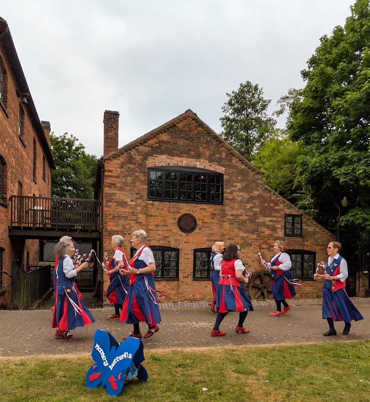 Nancy Butterfly dancing in front of the old Needle Mill, with their butterfly shaped blue sign on the grass in front of them