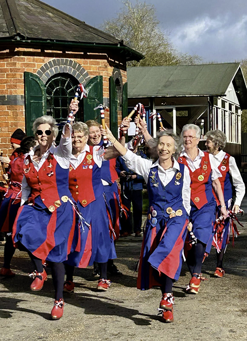 6 happy dancers in column of pairs moving towards camera with sticks raised