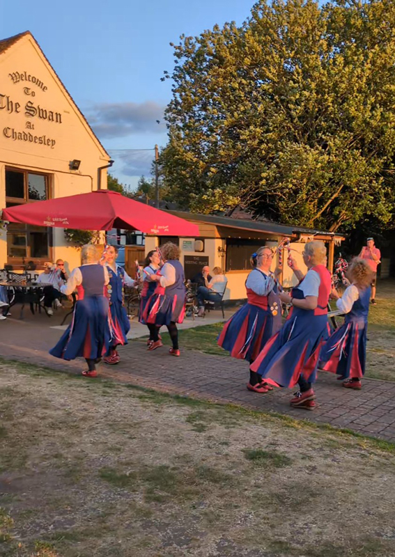 View from the side of Nancy Butterfly dancing on the path in the garden of the Swan Inn whose name features on the wall behind. There is also a large red sunshade adjacent