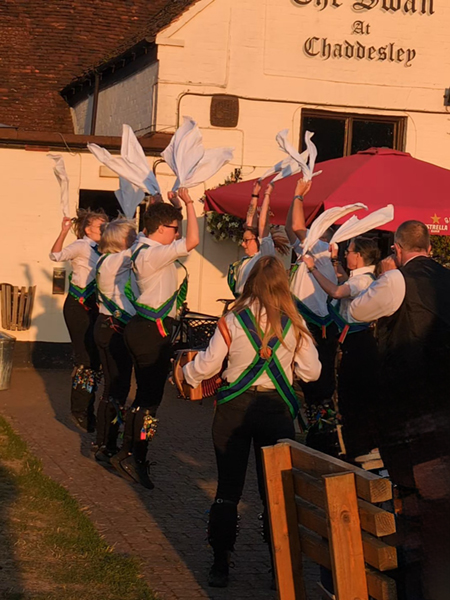 Bedcote Morris in green baldricks, white shirts and black breeches performing a dance using white handkerchiefs in front of 2 musicians