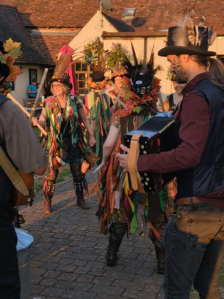 Bow Brook dancers in their colourful tatter jackets and feathered head dress, dancing, framed between 2 of their musicians