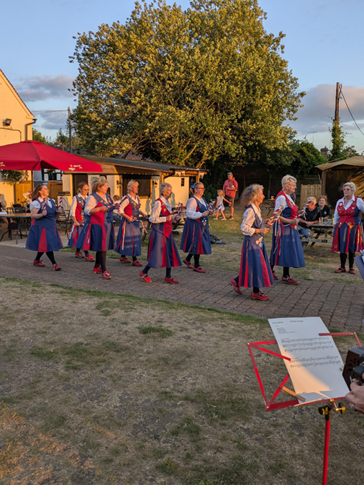 8 members of Nancy Butterfly, arranged in pairs and carrying short decorated sticks with bells, performing a dance along the path