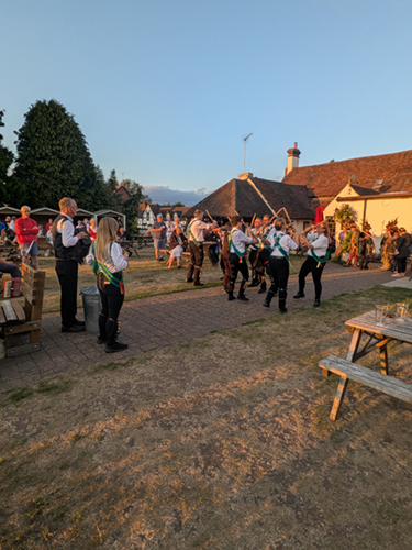 wide view of the garden with Bedcote Morris dancers and musicians performing in front of several spectators, all illuminated by a golden glow from a setting sun 