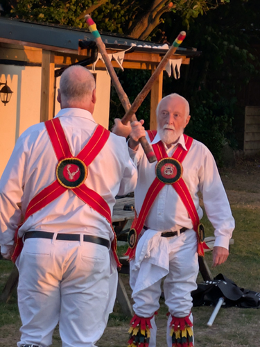 2 members of White Hart Morris in red baldricks, white shirt and trousers dancing in cotswold style with long sticks.