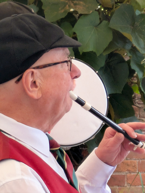 Head shot of a White Hart musician playing a traditional pipe, large green leaves in the background.