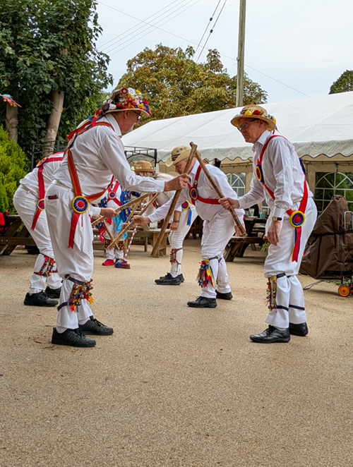 Gloucestershire Morris in their traditional Cotswold attire, including straw hats, performinhg a dance with clashing wooden sticks, on a hard standing area in the garden of the Old Bull in Inkberrow.